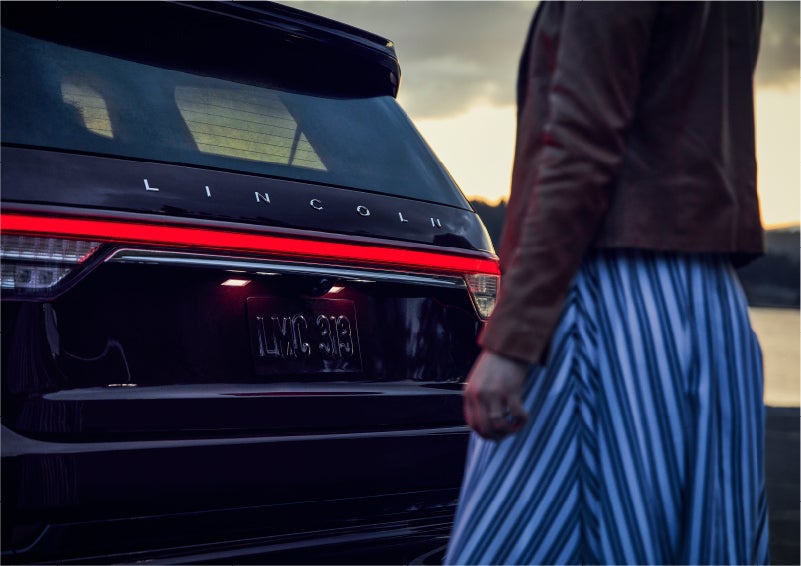 A person is shown near the rear of a 2023 Lincoln Aviator® SUV as the Lincoln Embrace illuminates the rear lights | Rogers Lincoln in Midland TX