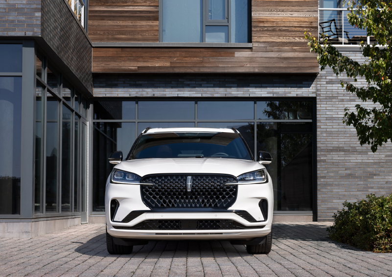 A white 2025 Lincoln Aviator® SUV is shown parked outside a modern building with gray brick and wood accents. | Rogers Lincoln in Midland TX