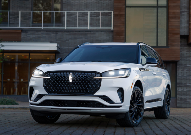 A white 2025 Lincoln Aviator® SUV is shown parked outside a modern building with gray brick and wood accents. | Rogers Lincoln in Midland TX